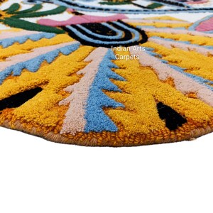 May include: Close-up of a round, textured rug with a colorful, abstract design. The rug features a yellow background with blue, pink, and black accents. The text "Indian Arts Carpets" is visible on the rug.