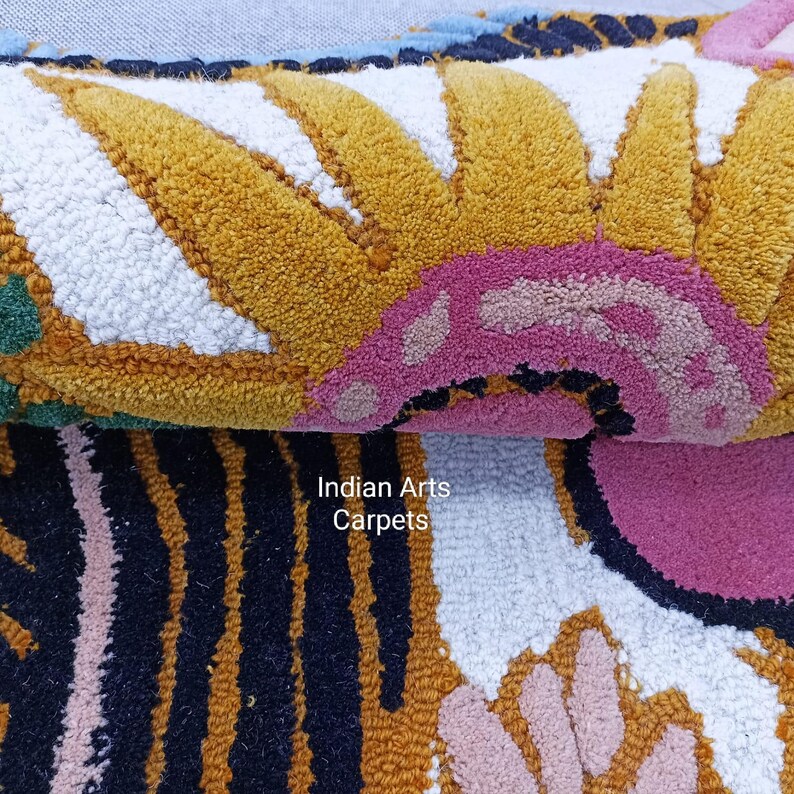 May include: Close-up of a colorful, textured rug with a floral design. The rug features a repeating pattern of yellow, white, pink, and black shapes. The text "Indian Arts Carpets" is visible in the lower right corner.