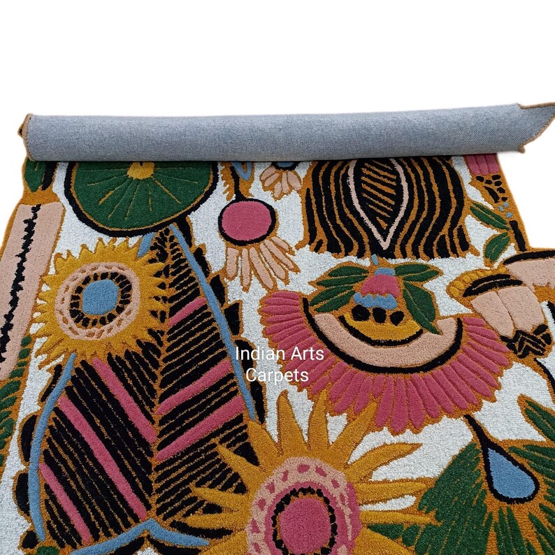 May include: A rolled-up, colorful rug with a floral design. The rug features a white background with large, stylized flowers in shades of pink, yellow, green, and black. The text "Indian Arts Carpets" is visible on the rug.