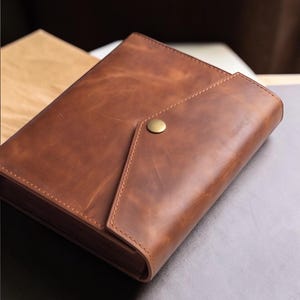May include: A brown leather journal with a flap closure and a brass button. The journal has a textured surface and appears to be new. The cover is a rich, warm brown color.
