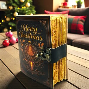 May include: A vintage-style journal with a black cover featuring the words "Merry Christmas" in gold script, along with a Christmas tree and candle illustration. The journal has aged, yellowed pages and a black leather strap closure. The journal is on a wooden surface.