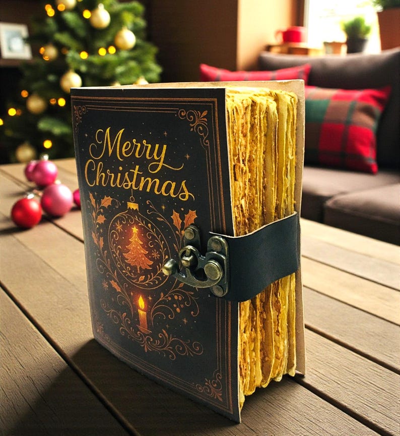 May include: A vintage-style journal with a black cover featuring the words "Merry Christmas" in gold script, along with a Christmas tree and candle illustration. The journal has aged, yellowed pages and a black leather strap closure. The journal is on a wooden surface.