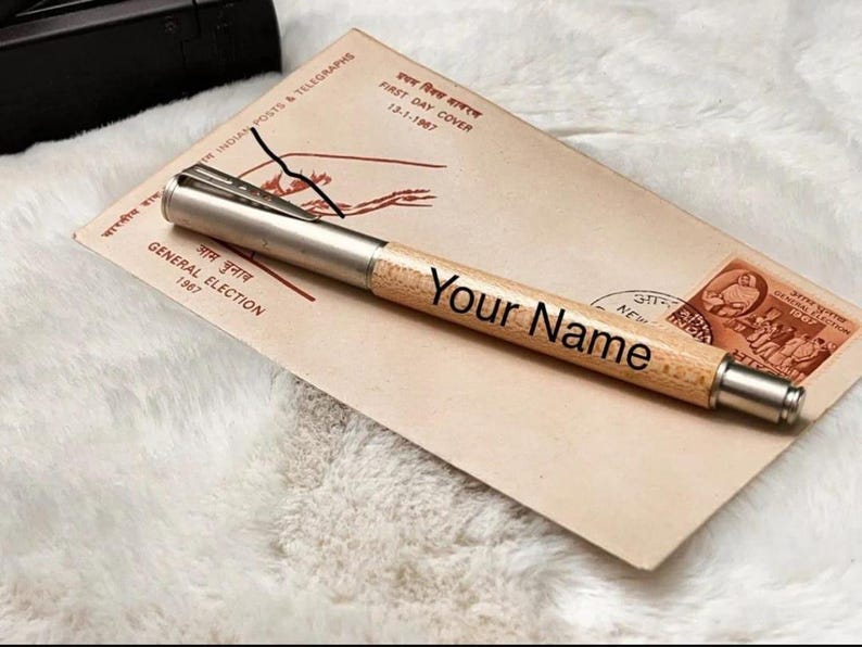May include: A personalized wooden pen with a silver metal tip and clip, engraved with the text "Your Name." The pen rests on a vintage envelope with text and stamps. The envelope is a light tan color.