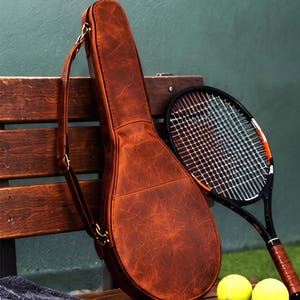 Pode incluir: Uma bolsa de couro castanho para raquete de ténis com acabamento texturizado e alça ajustável, acompanhada por uma raquete de ténis preta com cordas brancas e duas bolas de ténis amarelo brilhante sobre uma superfície de madeira.