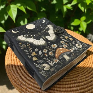 Puede incluir: Diario de cuero negro con diseño de mariposa y flores. La portada presenta una gran mariposa con alas blancas y negras, rodeada de margaritas, setas y lunas crecientes. El diario tiene un cierre metálico.