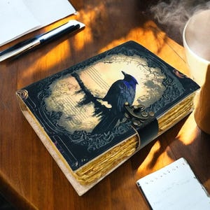 May include: A leather-bound journal with a raven illustration on the cover. The cover features a dark blue and black raven perched on a branch against a muted background. The journal is secured with a black leather strap and metal clasp.