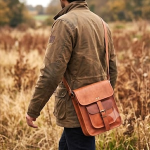 May include: A tan leather messenger bag with a front flap, a buckle closure, and a long shoulder strap. The bag features a rectangular main compartment and a smaller front pocket, worn over a brown jacket with elbow patches and a high collar.
