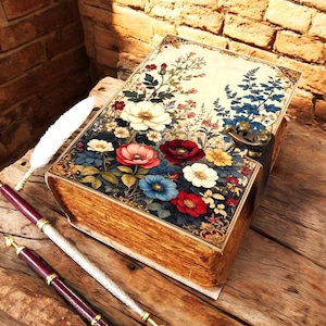 Könnte beinhalten: Eine rechteckige Box im Design eines alten Buches mit einem Blumenmuster in Rot, Weiß, Blau und Gelb auf dem Deckel. Die Box verfügt über einen messingfarbenen Metallverschluss an der Seite und wird von einer weißen Federkiel und zwei Stiften begleitet.