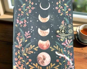 以下が含まれることがあります： 月の満ち欠け、星、花のアクセントが特徴の、暗い色の天体デザインのジャーナル。 カバーには金属製の留め金が付いています。 ジャーナルは、木製のテーブルの上に置かれ、お茶のカップが添えられています。