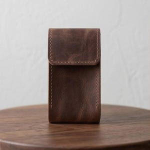 Może przedstawiać: Brązowe skórzane etui z klapką. Prostokątne etui ma widoczne szwy wzdłuż krawędzi. Skóra ma postarzany wygląd, a etui leży na drewnianej powierzchni.