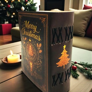 May include: A decorative Christmas book with the words "Merry Christmas" in gold script. The cover features a gold Christmas tree and ornament design. The spine has black stitching and a gold Christmas tree. A lit candle sits nearby.