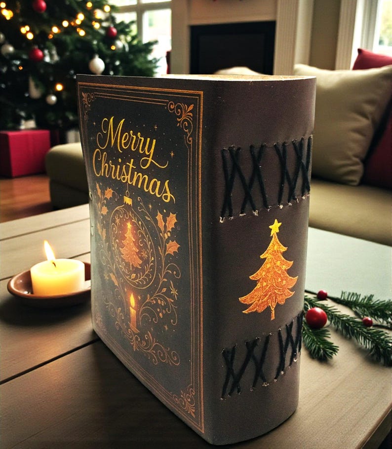 May include: A decorative Christmas book with the words "Merry Christmas" in gold script. The cover features a gold Christmas tree and ornament design. The spine has black stitching and a gold Christmas tree. A lit candle sits nearby.
