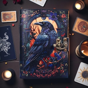 Peut inclure: Un journal en cuir bleu foncé avec une illustration détaillée d'un corbeau devant une fenêtre cintrée. La couverture présente des motifs floraux et un fermoir en métal. Cartes de tarot et bougies entourent le journal.