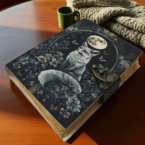 Könnte beinhalten: Ein dekoratives Notizbuch mit einer Fuchsdarstellung auf dem Einband. Der Einband zeigt einen Fuchs unter einem Vollmond, umgeben von Blumen und Schmetterlingen. Das Notizbuch hat einen Metallverschluss und einen dunkelblauen Einband.