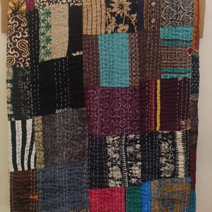 May include: A patchwork quilt with a variety of patterned fabrics in shades of brown, black, blue, and pink. The quilt is hand-stitched with white thread, creating a textured surface. The quilt is displayed on a wooden frame.