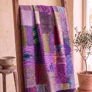 May include: A patchwork quilt in shades of purple, lavender, and magenta is draped over a wooden ladder. The quilt features a variety of patterned fabric squares, with visible stitching. The ladder is made of light brown wood and has carved details.