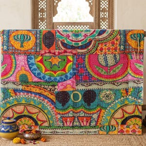 May include: A brightly coloured patchwork quilt, displayed on a wooden rack. The quilt features a variety of patterns in shades of pink, blue, yellow, and green. Decorative items, including a blue and yellow jar and a silver bowl, are placed in front.