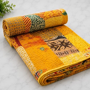 May include: A folded patchwork quilt featuring a vibrant yellow base colour with various rectangular panels displaying intricate patterns in shades of green, brown, and orange. The fabric surface shows detailed stitching throughout the entire textile.