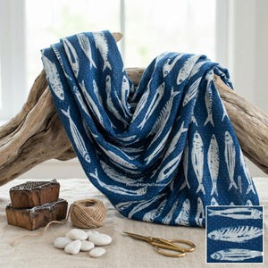 May include: A blue and white fish-print textile draped over driftwood. The fabric features a repeating pattern of white fish on a dark blue background. Additional items include wooden printing blocks, twine, white stones, and gold scissors.