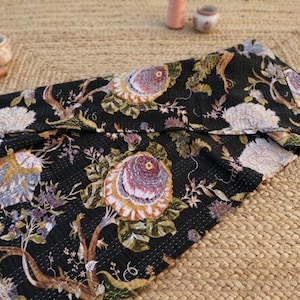 Può includere: Tessuto nero con motivo floreale con grandi fiori stilizzati nei toni del viola, blu e crema. Il tessuto è piegato, rivelando l'intricato design. Lo sfondo è un tappeto in fibra naturale intrecciata.