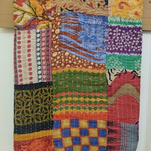 May include: A patchwork textile with a variety of patterned squares in shades of blue, red, green, yellow, and brown. The textile is stitched together with white thread. The textile is hanging from a wooden bar.