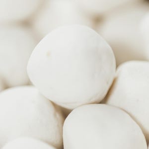 Op de afbeelding: Een close-up van een stapel witte, ronde voedingsmiddelen, mogelijk kaas- of deegballen. De items zijn dicht op elkaar gepakt, met een zachte, vage achtergrond. Het logo van Mama Sasi, The Modern Greek Deli, staat rechtsonder.