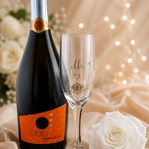 May include: A bottle of Prosecco with an orange label and a champagne flute with the name "Dulhan" and a lotus flower design. A white rose and decorative pearls are also present.