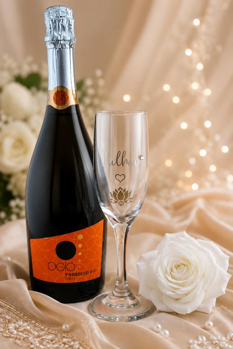 May include: A bottle of Prosecco with an orange label and a champagne flute with the name "Dulhan" and a lotus flower design. A white rose and decorative pearls are also present.
