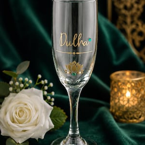 May include: A clear champagne flute with the word "Dulha" in gold script. The glass is decorated with a gold lotus flower and small gemstones. The background is a dark green velvet.