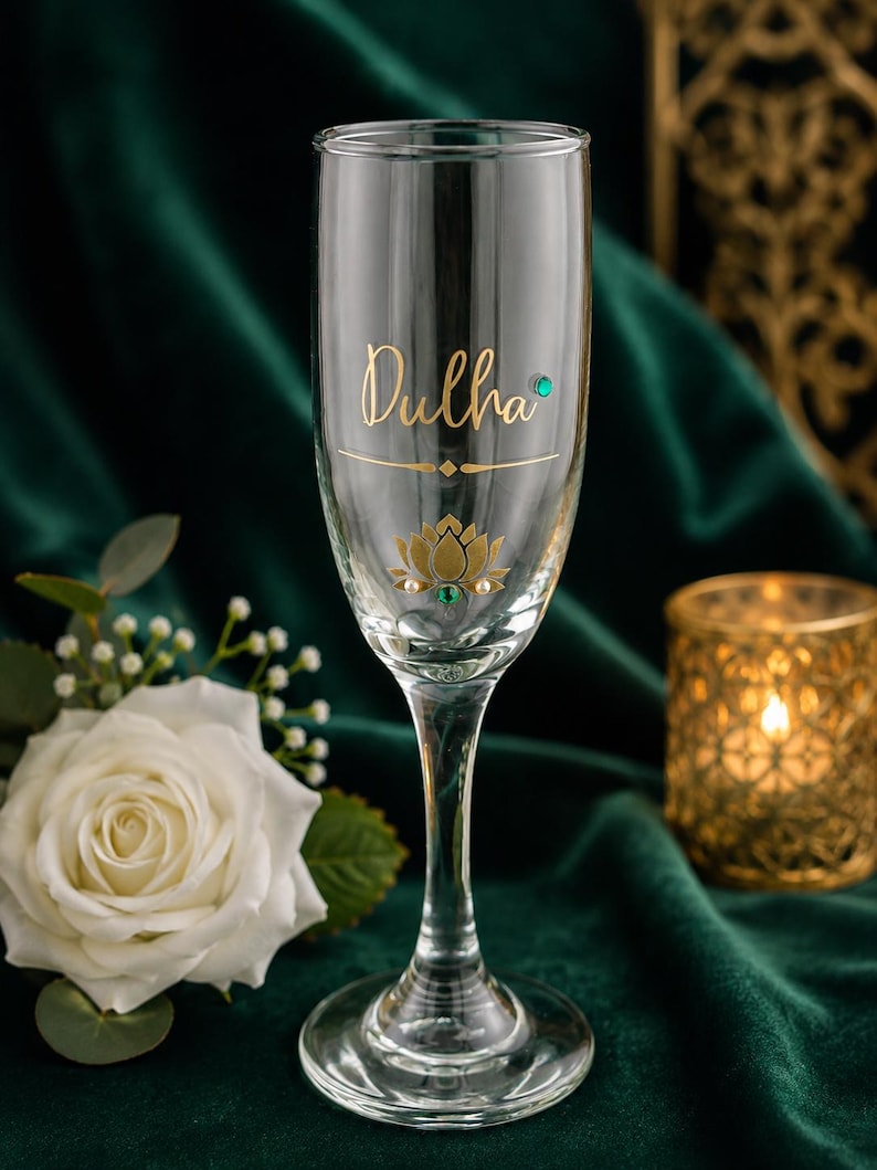 May include: A clear champagne flute with the word "Dulha" in gold script. The glass is decorated with a gold lotus flower and small gemstones. The background is a dark green velvet.