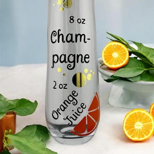May include: A clear champagne flute with black text that reads "8 oz Champagne 2 oz Orange Juice." The glass has a decorative orange slice graphic and gold bubble accents. The glass is next to orange slices and green leaves.