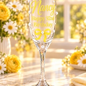 May include: A clear glass champagne flute decorated with white pearl accents and a yellow ribbon graphic. The glass features the yellow text Nancy Happy 92nd Birthday printed on the front surface, with a slender stem and a circular base.