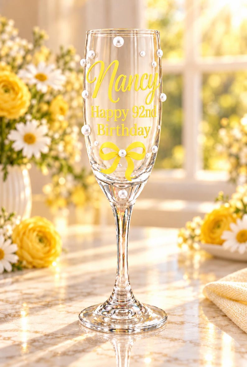 May include: A clear glass champagne flute decorated with white pearl accents and a yellow ribbon graphic. The glass features the yellow text Nancy Happy 92nd Birthday printed on the front surface, with a slender stem and a circular base.