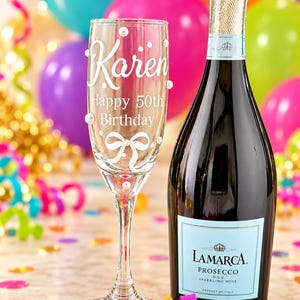 May include: A clear glass champagne flute featuring the white text Karen Happy 50th Birthday and a white bow graphic, accented with small white pearls, standing next to a dark glass bottle of La Marca Prosecco with a light blue label.