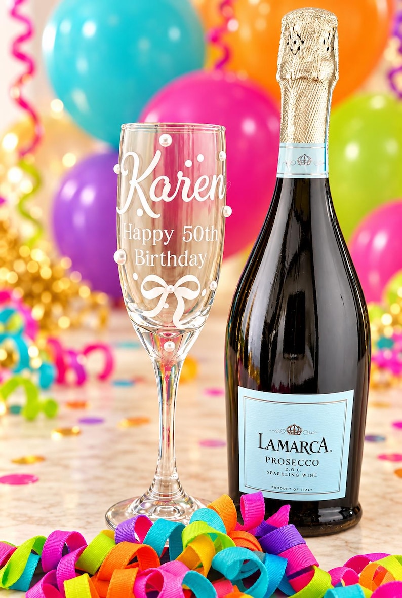 May include: A clear glass champagne flute featuring the white text Karen Happy 50th Birthday and a white bow graphic, accented with small white pearls, standing next to a dark glass bottle of La Marca Prosecco with a light blue label.