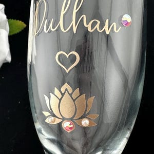 May include: A clear champagne flute with gold lettering that reads "Dulhan". The glass is decorated with a gold lotus flower, a heart, and iridescent and pearl-like embellishments. The glass is on a black background.