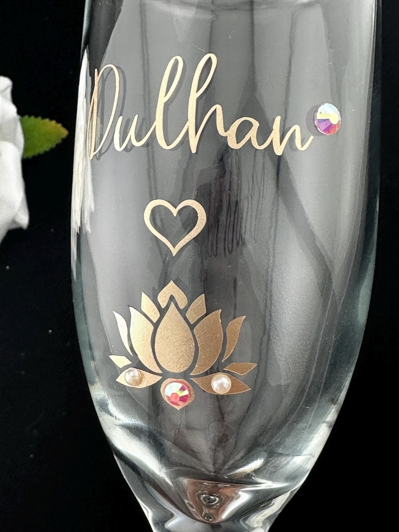 May include: A clear champagne flute with gold lettering that reads "Dulhan". The glass is decorated with a gold lotus flower, a heart, and iridescent and pearl-like embellishments. The glass is on a black background.