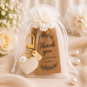 May include: A white organza drawstring favor bag topped with a white fabric flower. Inside is a gold-toned keychain with a heart-shaped charm covered in small white pearls and a rectangular tag printed with Thank you for celebrating with us.