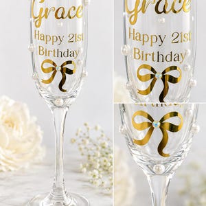 May include: A clear glass champagne flute featuring gold lettering that reads Grace Happy 21st Birthday. The glass is decorated with a gold foil bow graphic with a central iridescent crystal accent and several small white faux pearls attached to the exterior.