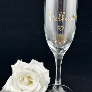 May include: A clear champagne flute with the word "Dulhan" in gold script. The glass is decorated with a gold lotus flower, heart, and gemstones. A white rose rests beside the glass against a black background.
