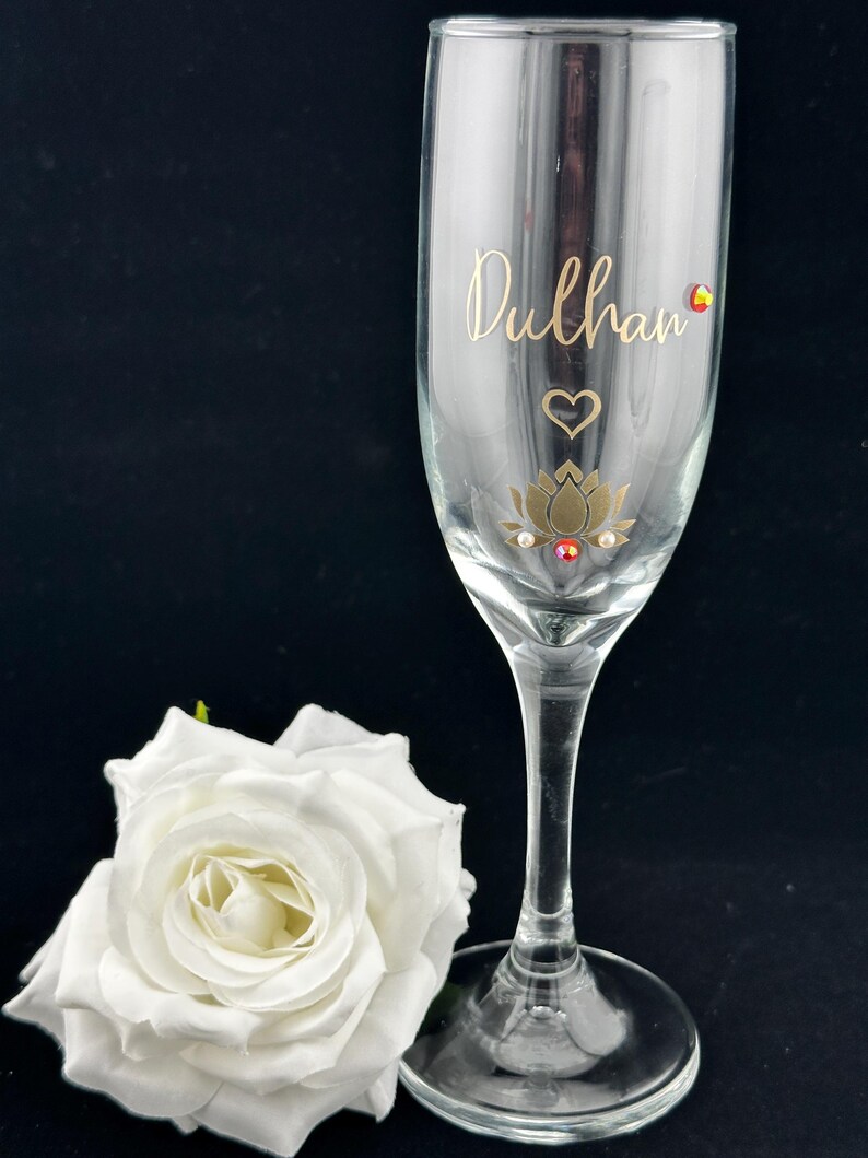 May include: A clear champagne flute with the word "Dulhan" in gold script. The glass is decorated with a gold lotus flower, heart, and gemstones. A white rose rests beside the glass against a black background.