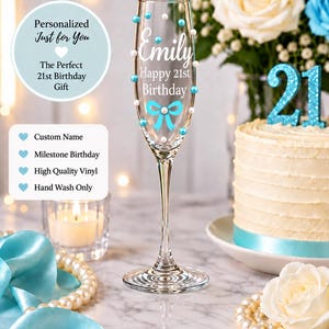 May include: A clear glass champagne flute decorated with white and light blue pearls, featuring the white text Emily, Happy 21st Birthday, and a light blue bow graphic. The glass has a slender stem and a circular base, standing upright.