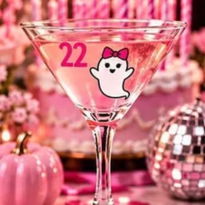 May include: A clear martini glass featuring a pink graphic of the number 22 and a white ghost wearing a pink bow. The glass has a stem base decorated with three small white pearls, and it is filled with a light pink liquid beverage.
