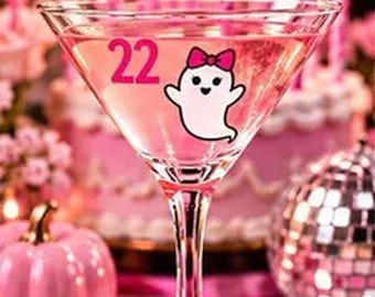 May include: A clear martini glass featuring a pink graphic of the number 22 and a white ghost wearing a pink bow. The glass has a stem base decorated with three small white pearls, and it is filled with a light pink liquid beverage.