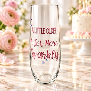 May include: A clear stemless champagne flute featuring the pink text A LITTLE OLDER A Lot More Sparkly. The glass is decorated with small, iridescent rhinestones scattered around the lettering, creating a reflective and decorative appearance.