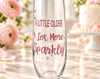 May include: A clear stemless champagne flute featuring the pink text A LITTLE OLDER A Lot More Sparkly. The glass is decorated with small, iridescent rhinestones scattered around the lettering, creating a reflective and decorative appearance.