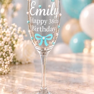 May include: A clear glass champagne flute featuring the white text Emily Happy 38th Birthday. The glass is decorated with small light blue and white pearl-like beads and a light blue bow graphic printed on the front surface of the bowl.