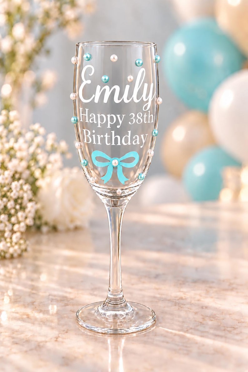 May include: A clear glass champagne flute featuring the white text Emily Happy 38th Birthday. The glass is decorated with small light blue and white pearl-like beads and a light blue bow graphic printed on the front surface of the bowl.
