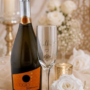 May include: A bottle of Ogio Prosecco DOC Italia next to a champagne flute with the name "Dullhan" and a lotus flower design. A white rose and a lit candle are also in the image.