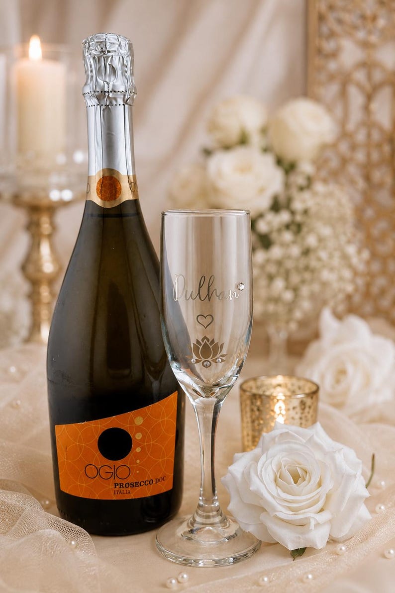 May include: A bottle of Ogio Prosecco DOC Italia next to a champagne flute with the name "Dullhan" and a lotus flower design. A white rose and a lit candle are also in the image.